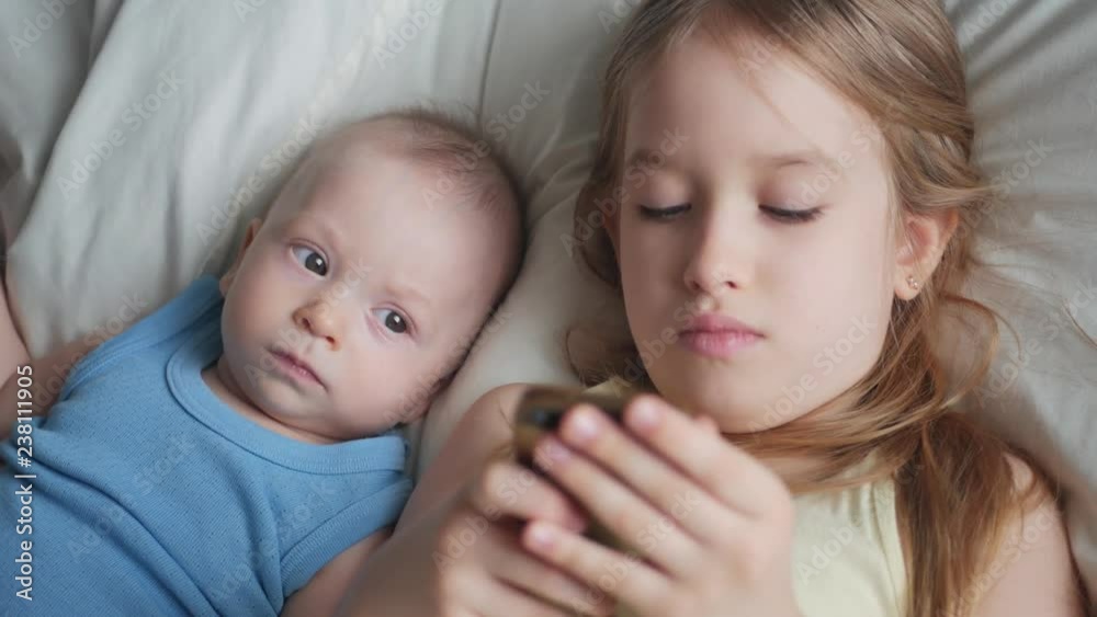 Two children use and play mobile phone together on their bed. Kids use telephone together on bed in the bedroom. Relationship of brother and sister concept. Two children use and play mobile phone together on their bed. Kids use telephone together on bed in the bedroom. Relationship of brother and sister concept.