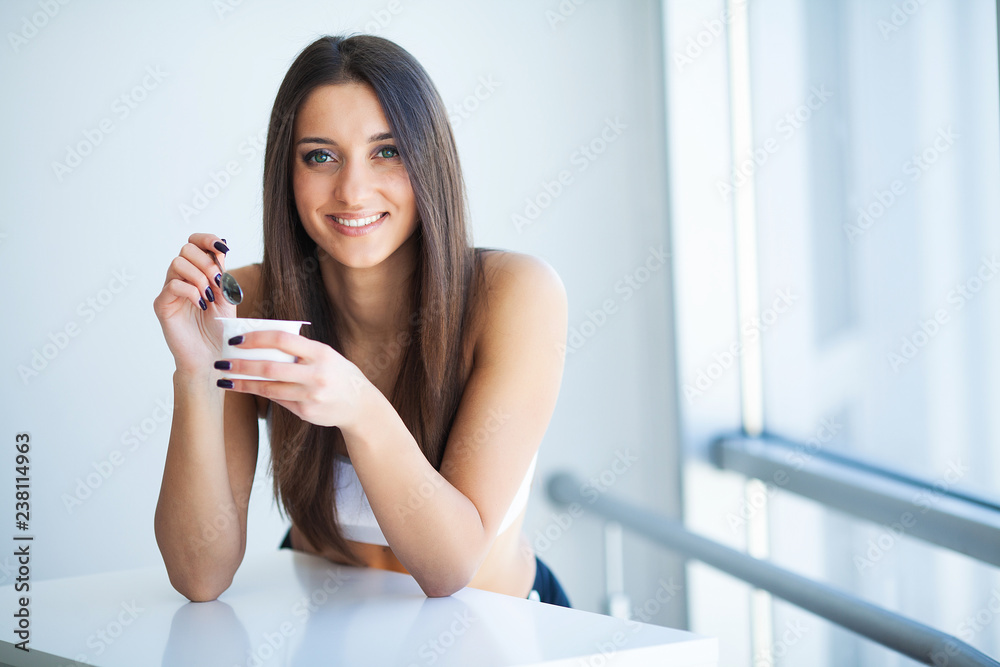 Smiling Girl With Yogurt. Young smiling Woman Tasting Fresh Organic Yogurt sitting in white bright room, wearing in white singlet. Healthy Lifestyle Concept
