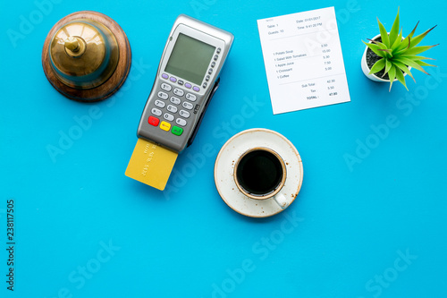 Electronic payments. Pay the bill by card concept. Bank card inserted in payment terminal near bill, service bell, coffee on blue background top view copy space