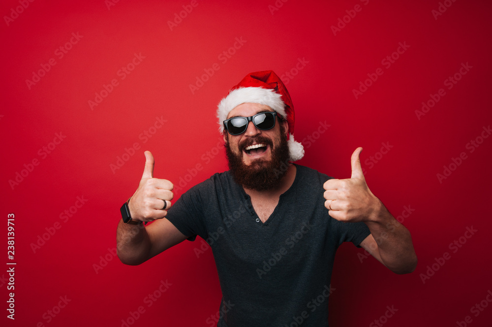 Smiling man with beard wearing santa claus hat showing thumbs up gesture over red background Smiling man with beard wearing santa claus hat showing thumbs up gesture over red background