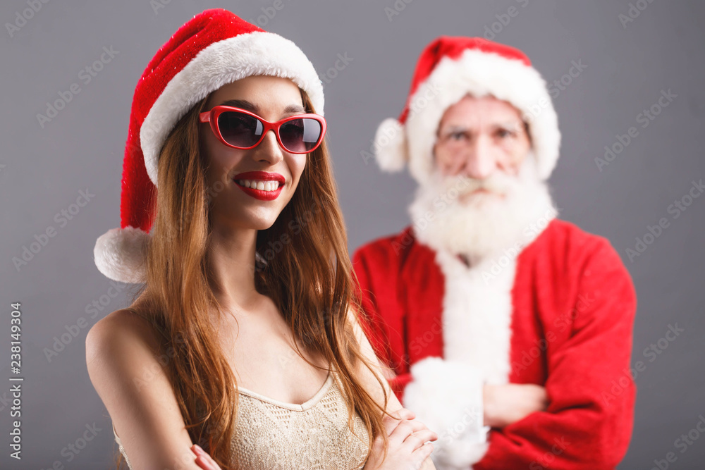 Young mrs. Claus wearing Santa hat and sunglasses standing and smiling, Santa Claus standing behind on the gray background, New Year, Christmas, holidays, souvenirs, gifts, shopping, discounts, shops
