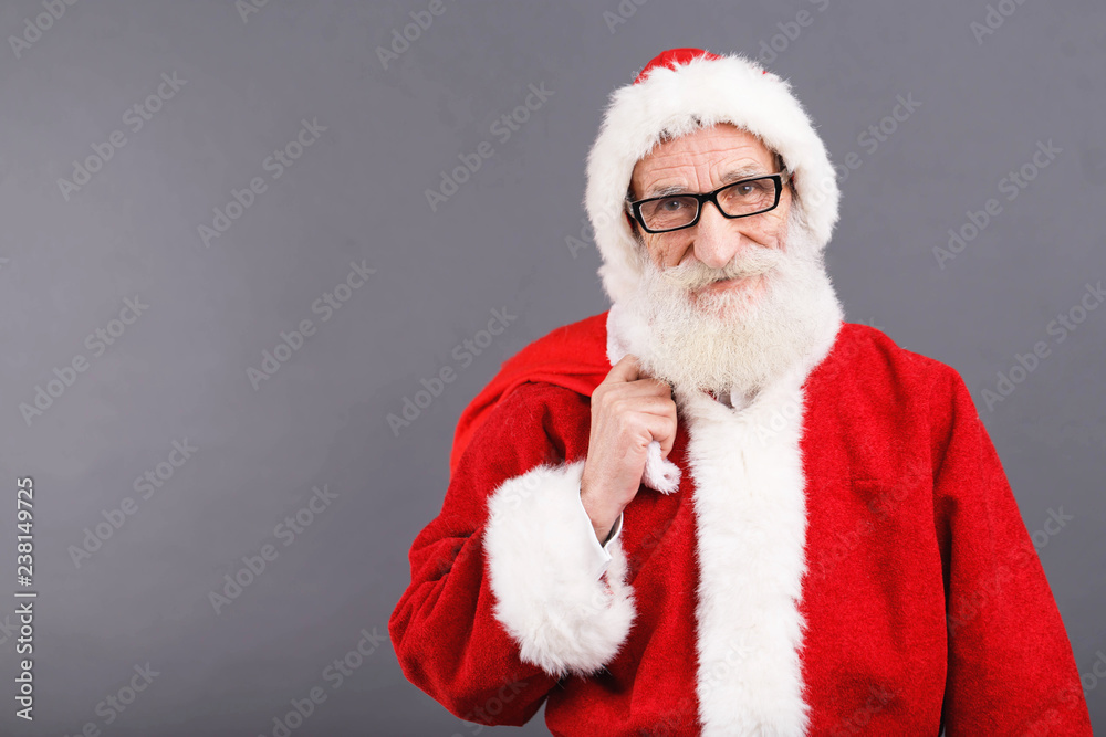 Santa Claus with a white beard wearing Santa outfit standing with the bag on the gray background, New Year, Christmas, holidays, souvenirs, gifts, shopping, discounts, shops Santa Claus with a white beard wearing Santa outfit standing with the bag on the gray background, New Year, Christmas, holidays, souvenirs, gifts, shopping, discounts, shops