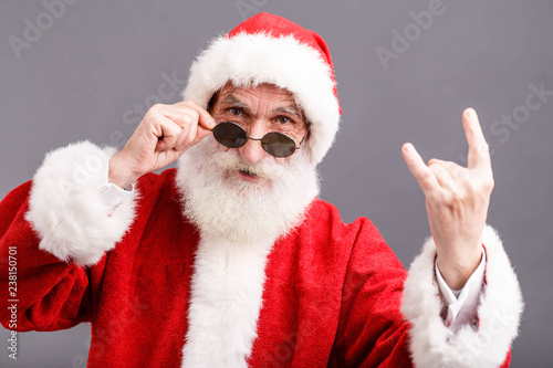 Portrait of Santa Claus with a white beard wearing sunglasses and Santa outfit standing and showing a rock gesture on the gray background  New Year  Christmas  holidays  souvenirs  gifts  shopping