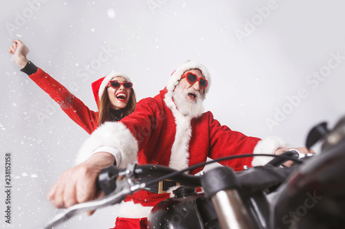 Santa Claus with white beard wearing sungasses and young mrs. Claus wearing Santa hat, red sweater and sunglasses rejoicing in the snow while riding a motorcycle when snowing, New Year, Christmas