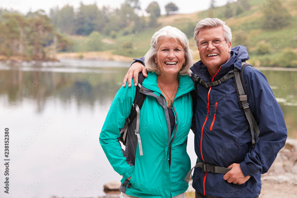 Happy senior couple standing on the shore of a lake smiling to camera, Lake District, UK Happy senior couple standing on the shore of a lake smiling to camera, Lake District, UK