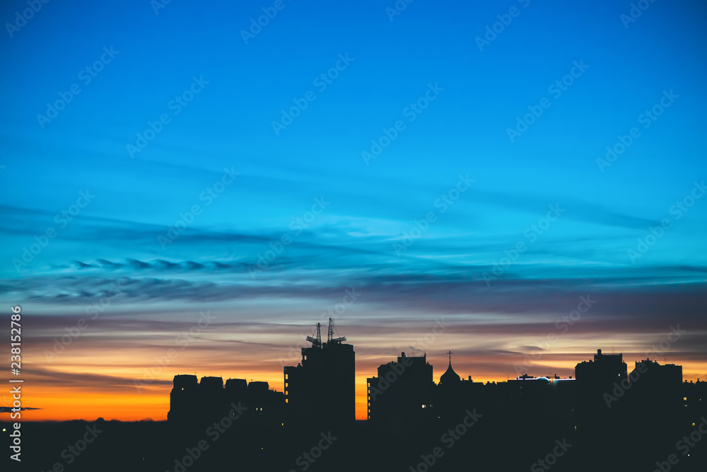 Cityscape with wonderful varicolored vivid dawn. Amazing dramatic blue sky with purple and violet clouds above dark silhouettes of city buildings. Atmospheric background of orange sunrise. Copy space.