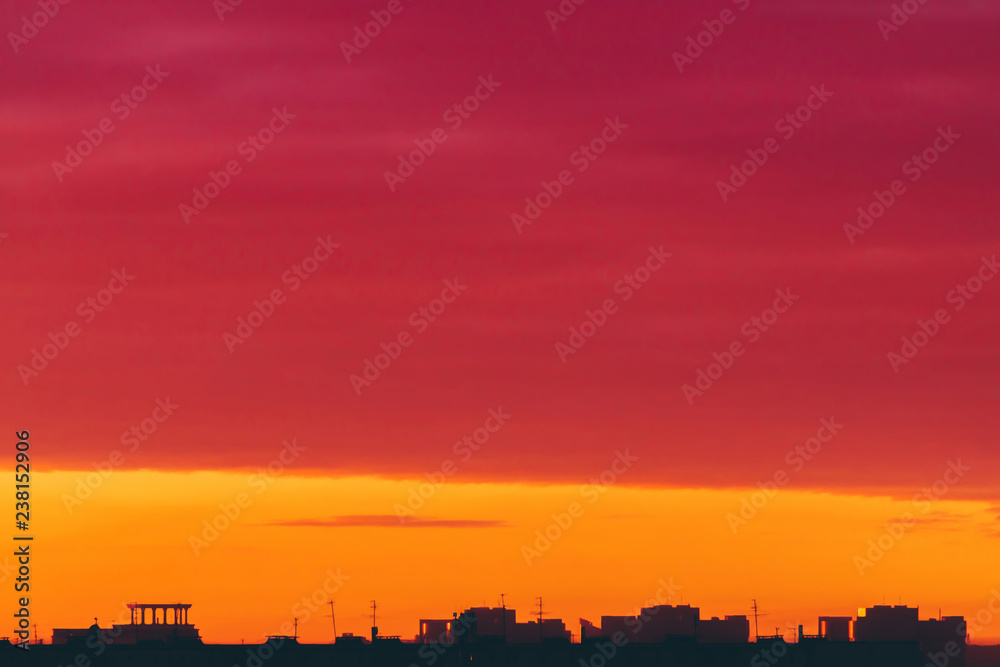 Cityscape with vivid surrealistic dawn. Amazing warm dramatic cloudy sky above dark silhouettes of city buildings. Orange sunlight. Atmospheric background of sunrise in overcast weather. Copy space.