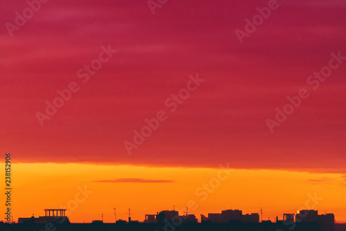 Cityscape with vivid surrealistic dawn. Amazing warm dramatic cloudy sky above dark silhouettes of city buildings. Orange sunlight. Atmospheric background of sunrise in overcast weather. Copy space.