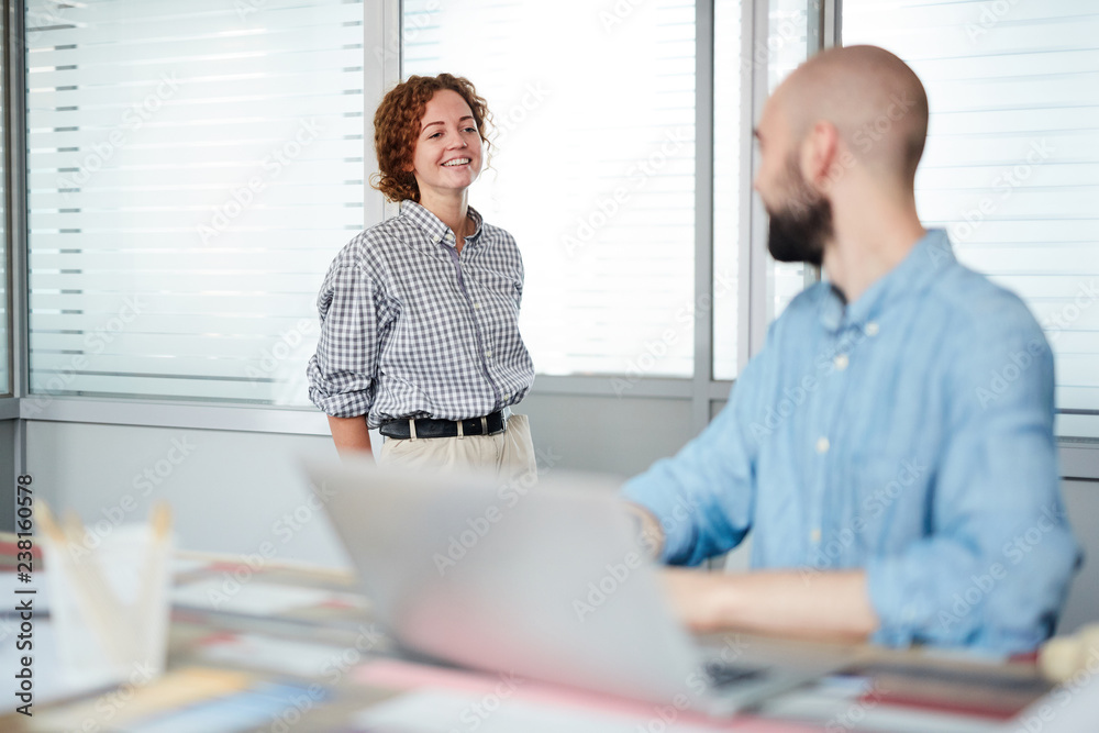 Obraz premium Smiling attractive lady designer with red hair leaning on doorjamb and talking to colleague typing on laptop in office
