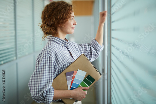 Smiling content confident beautiful design student holding sketchpad and color palettes and knocking on window while peeking through blinds in corridor