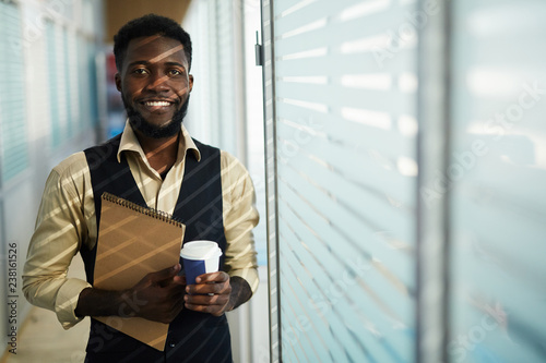 Smiling handsome young black designer with beard standing in narrow corridor of modern company and drinking coffee while looking at camera and holding sketchpad