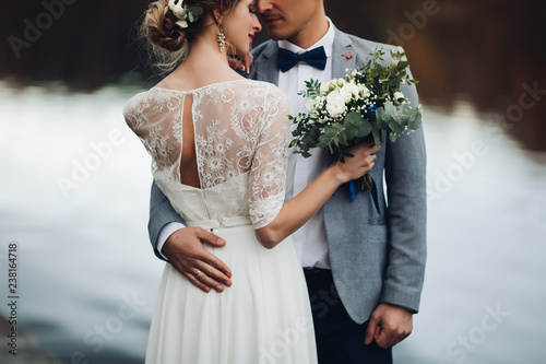 Back view of husband embracing his beautiful elegant wife in lace wedding dress with lovely bouquet against unfocused lake surface. photo