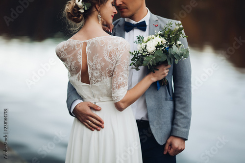 Back view of husband embracing his beautiful elegant wife in lace wedding dress with lovely bouquet against unfocused lake surface.