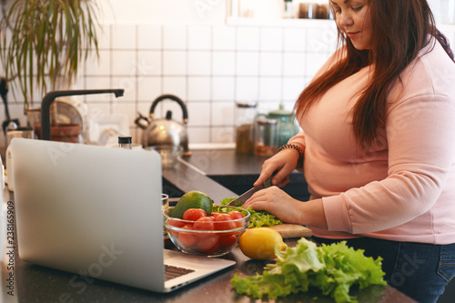 Overweight woman using laptop to watch video recipe while making vegan vitamin avocado salad, slicing leaf lettuce on wooden cutting board. Healthy food, weight loss, dieting and nutrition concept photo