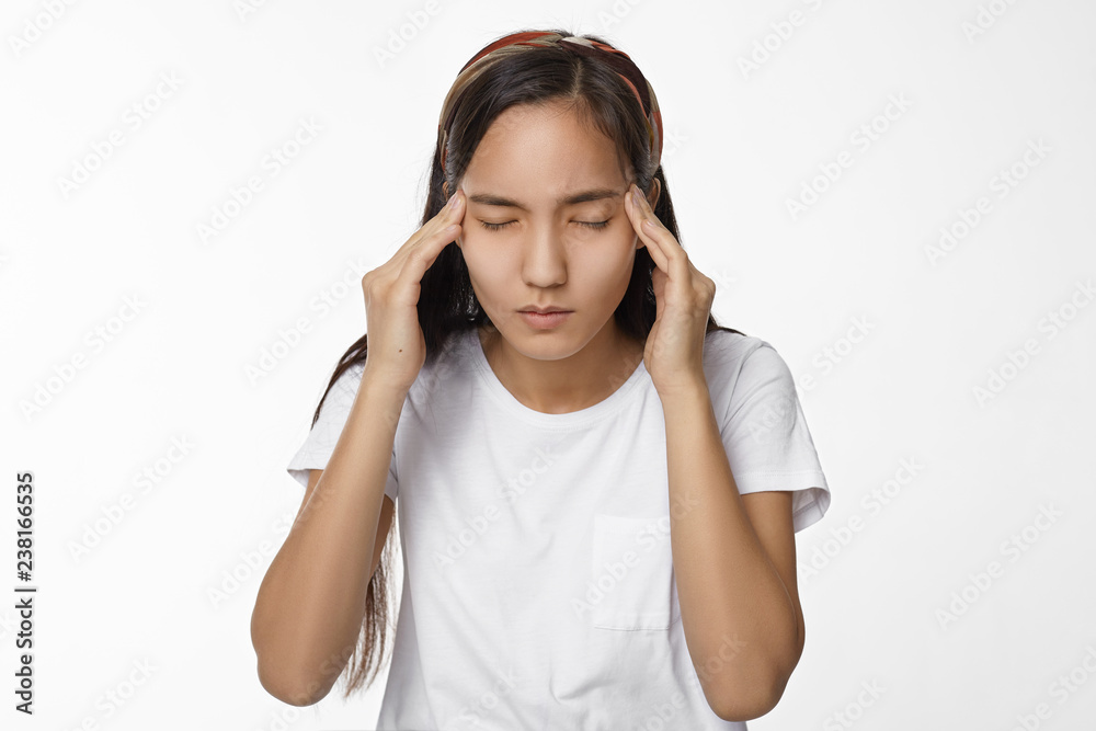 People, health and sickness concept. Brunette Asian student girl in white t-shirt closing eyes and squeezing her head, having bad headache, suffering from stress while preparing to important exams People, health and sickness concept. Brunette Asian student girl in white t-shirt closing eyes and squeezing her head, having bad headache, suffering from stress while preparing to important exams