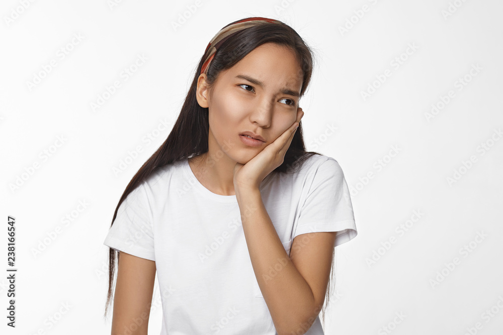 Isolated shot of dark haired frustrated young Chinese female having upset painful facial expression, holding hand on her cheek, suffering from toothache, posing isolated at white studio wall Isolated shot of dark haired frustrated young Chinese female having upset painful facial expression, holding hand on her cheek, suffering from toothache, posing isolated at white studio wall