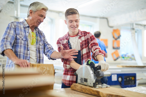 Positive young carpentry student in work gloves standing at workbench and using circular saw under control of experienced master at class in workshop