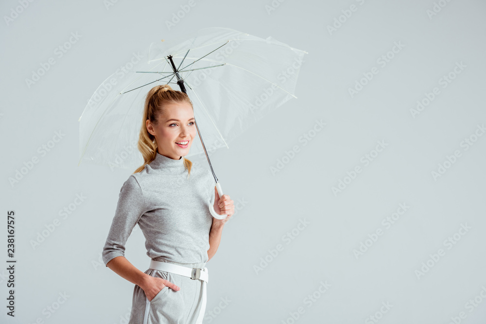 beautiful woman in grey clothes and hand in pocket posing with transparent umbrella isolated on grey - obrazy, fototapety, plakaty 
