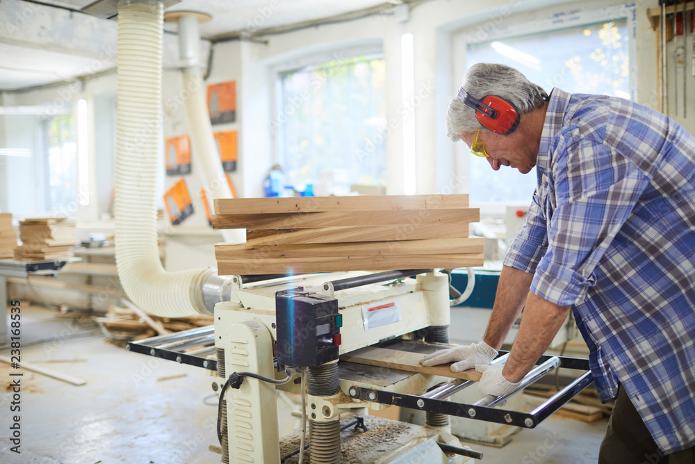 Serious concentrated senior carpenter with gray hair wearing ear protectors and safety goggles pushing wooden detail into machine and adjusting it in workshop