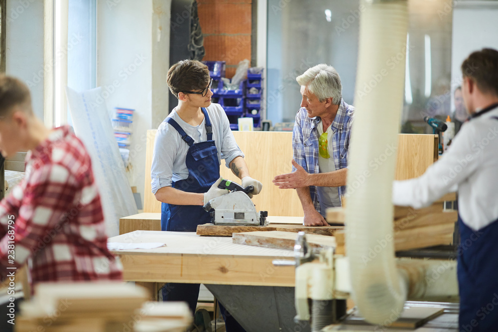 Serious skilled senior carpenter with gray hair standing at desk and gesturing hand while teaching young worker to use polish machine at factory