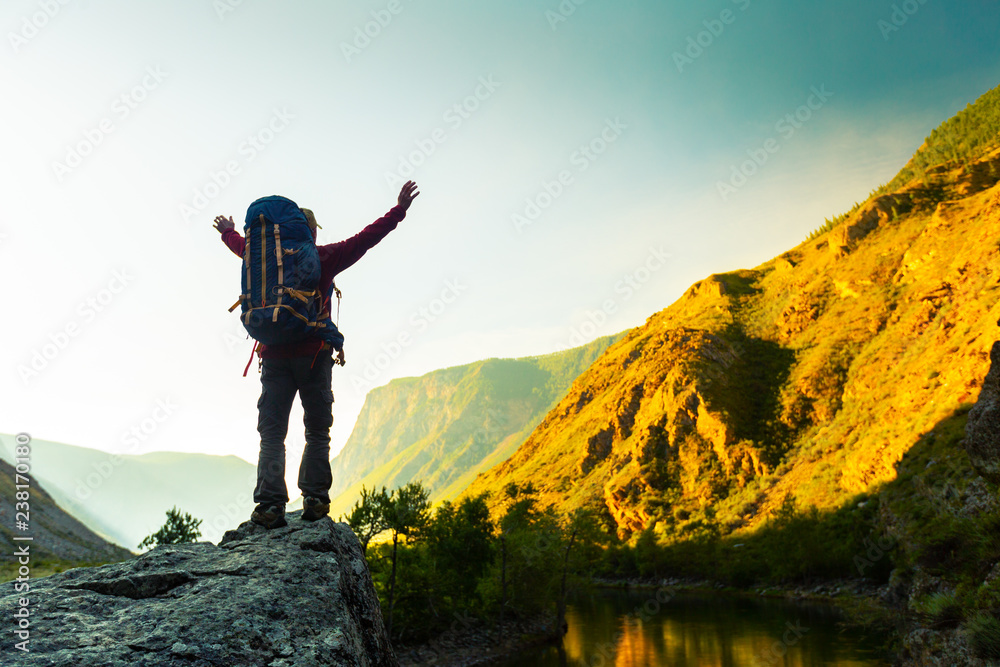 Hiker with backpack stands on the rock with raised hands and enjoys sunrise valley views. Altai, Russia