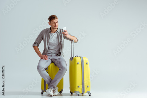 handsome man sitting on yellow suitcase and using smartphone on grey background, travel concept