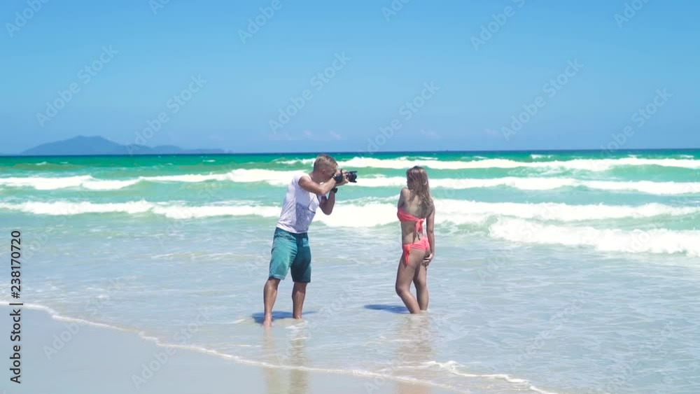Man photographer taking photography of young woman in bikini posing on sea beach. Young man photographer working with woman model on sea. Man using photo camera for photo session of beautiful woman. Man photographer taking photography of young woman in bikini posing on sea beach. Young man photographer working with woman model on sea. Man using photo camera for photo session of beautiful woman.
