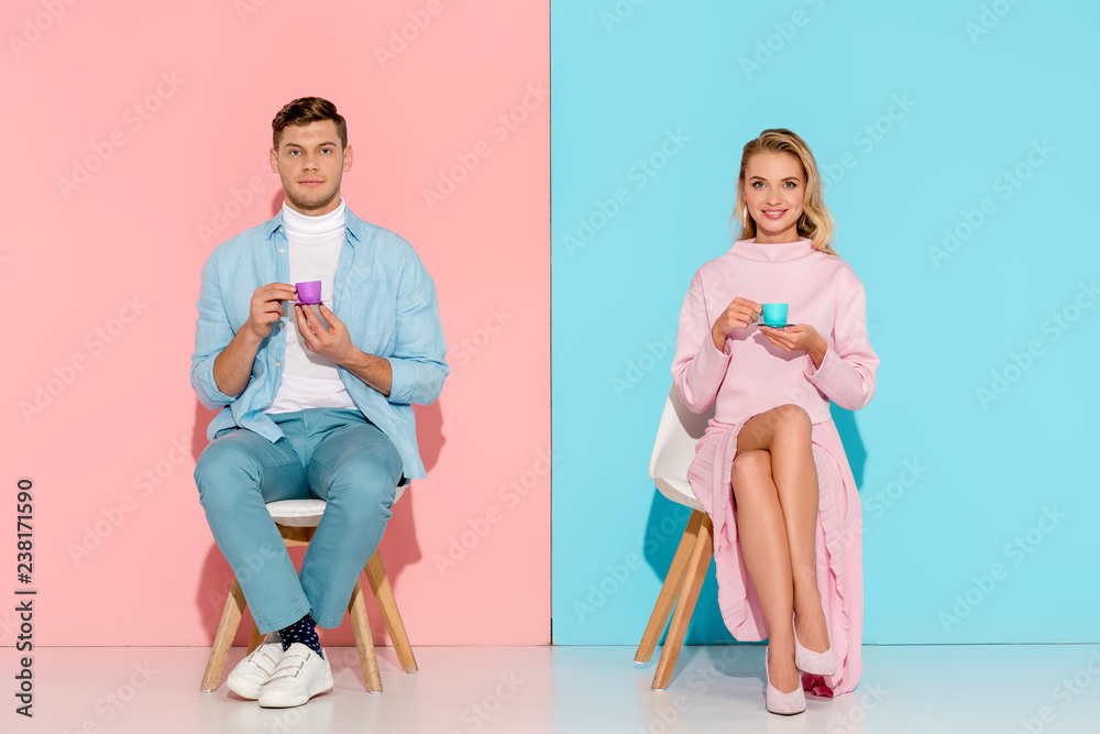 Obraz premium couple sitting on chairs, holding toy coffee cups and looking at camera on pink and blue background