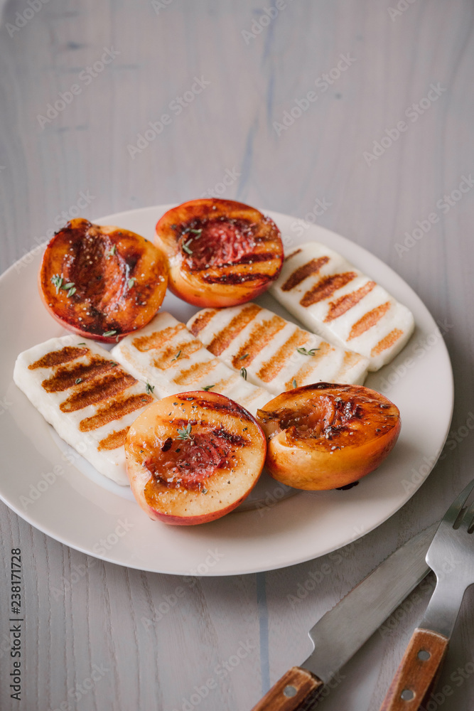 Grilled slices of halloumi cheese served on the plate with grilled peaches thyme leaf and balsamic. Halloumi is popular in Cyprus, Greece, and Turkey. Cutlery on a light grey background. Grilled slices of halloumi cheese served on the plate with grilled peaches thyme leaf and balsamic. Halloumi is popular in Cyprus, Greece, and Turkey. Cutlery on a light grey background.