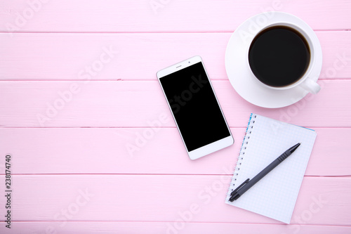 Smart phone with notebook and cup of coffee on pink wooden background