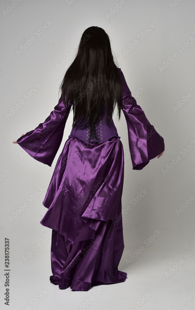 full length portrait of beautiful girl with long black hair, wearing purple fantasy medieval gown. standing pose with back to the camera on grey studio background. full length portrait of beautiful girl with long black hair, wearing purple fantasy medieval gown. standing pose with back to the camera on grey studio background.
