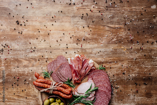 top view of cutting board with olives and sliced salami, prosciutto and ham on wooden table with scattered spices
