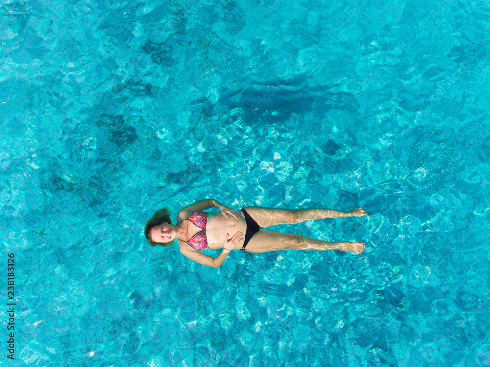 Aerial view of pregnant woman floating on her back in transparent sea.