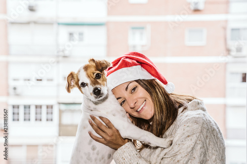 .Sweet woman enjoying with her nice dog jack russell. Looking out the window at the cold Christmas morning. She is wearing a santa claus hat. Lifestyle