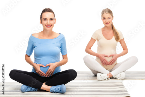 two beautiful pregnant women holding hands in heart sign on bellies isolated on white