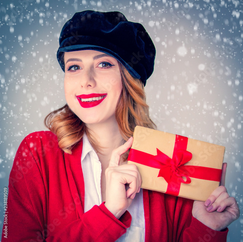 Portrait of young redhead woman in red cardigan and hat with gift box on grey background. Image made with native lights photo