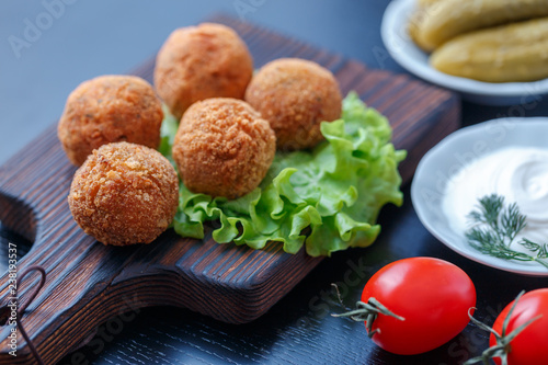 Falafel lies on a wooden cutting board. On the table lie tomatoes, cucumbers, lettuce, dill, lemon, sour cream.