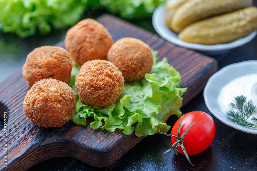 Falafel lies on a wooden cutting board. On the table lie tomatoes, cucumbers, lettuce, dill, lemon, sour cream.