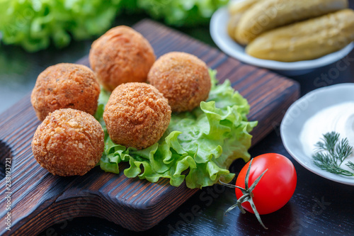 Falafel lies on a wooden cutting board. On the table lie tomatoes, cucumbers, lettuce, dill, lemon, sour cream.