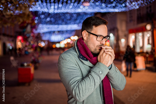 Winter portrait of handsome man trying to warm up his hands at night in the city street.