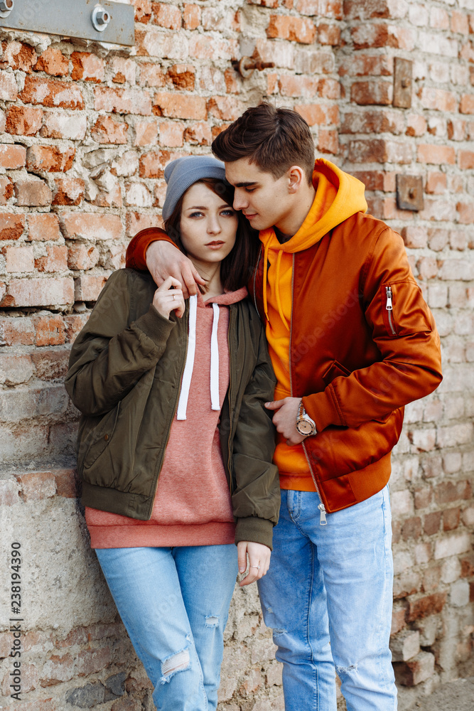 Young lovers hugging on the background of urban scenery. A guy and a girl in jeans and jackets posing against a brick wall.