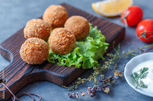 Falafel lies on a wooden cutting board. On the table lie tomatoes, cucumbers, lettuce, salad, dill, lemon, sour cream.