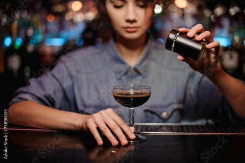Female bartender adding to a cocktail with brown alcohol a powder on the bar counter