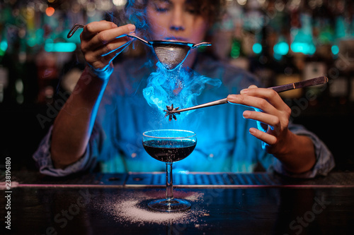 Girl adding to a brown cocktail and pour on a flamed badian on tweezers a powdered sugar through the strainer in the blue light photo