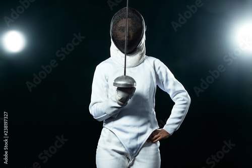 Young fencer athlete wearing fencing costume holding the sword and mask. Isolated on black background with lights photo