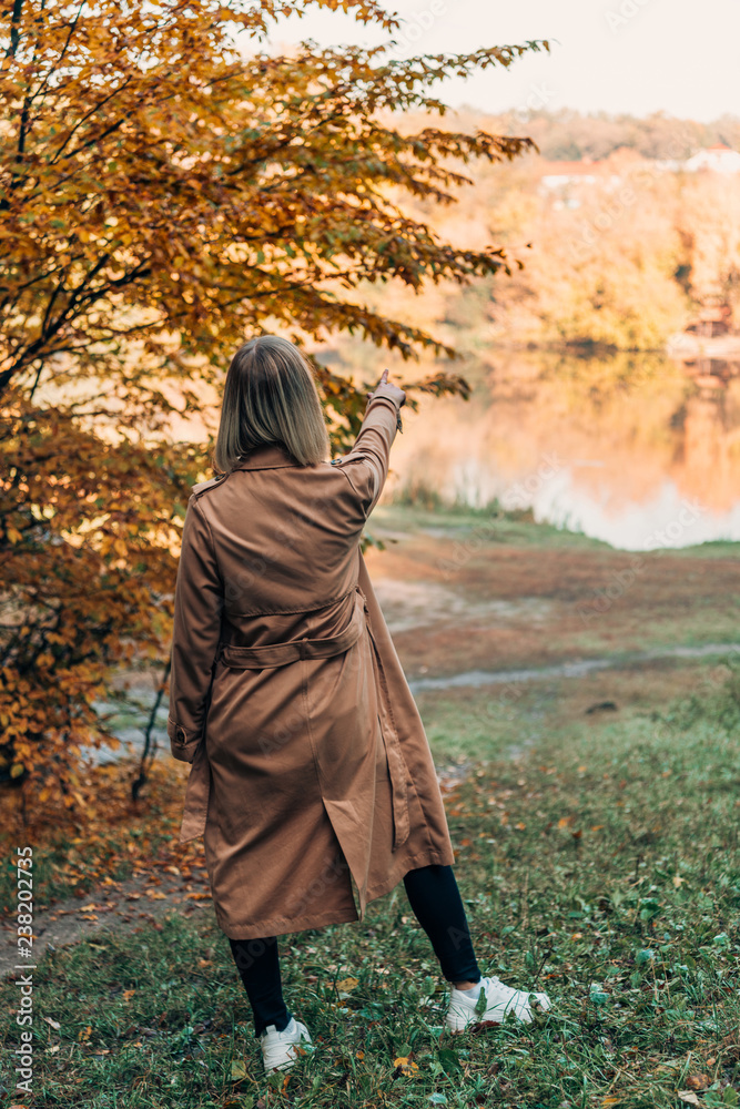 Back view of woman standing in coat and pointing at lake in autumn forest
