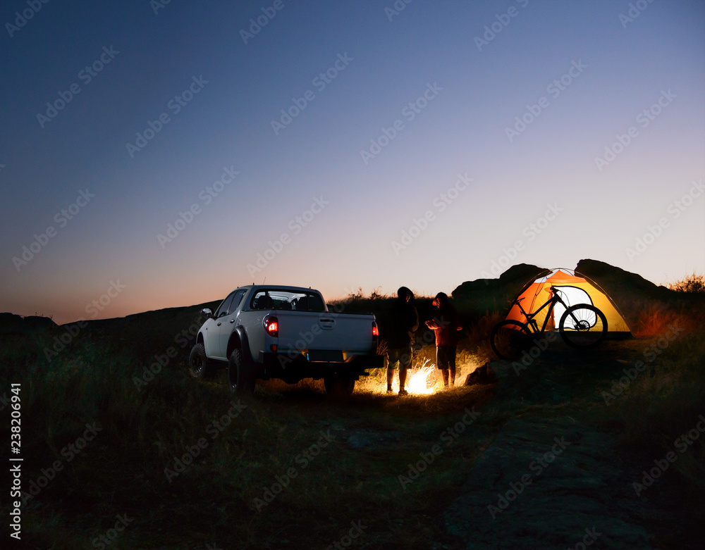 Friends near Bonfire, Pickup Truck, Tent and Bike at Night Camp in the Mountains. Adventure and Travel Concept - obrazy, fototapety, plakaty 