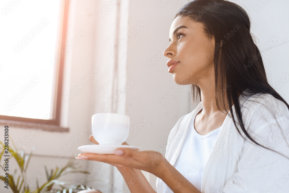 side view of beautiful mixed race girl in white robe holding cup of coffee in morning in bedroom with sunlight