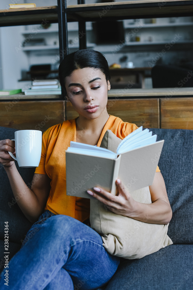 beautiful mixed race girl in orange shirt holding cup of tea and reading book on sofa at home beautiful mixed race girl in orange shirt holding cup of tea and reading book on sofa at home