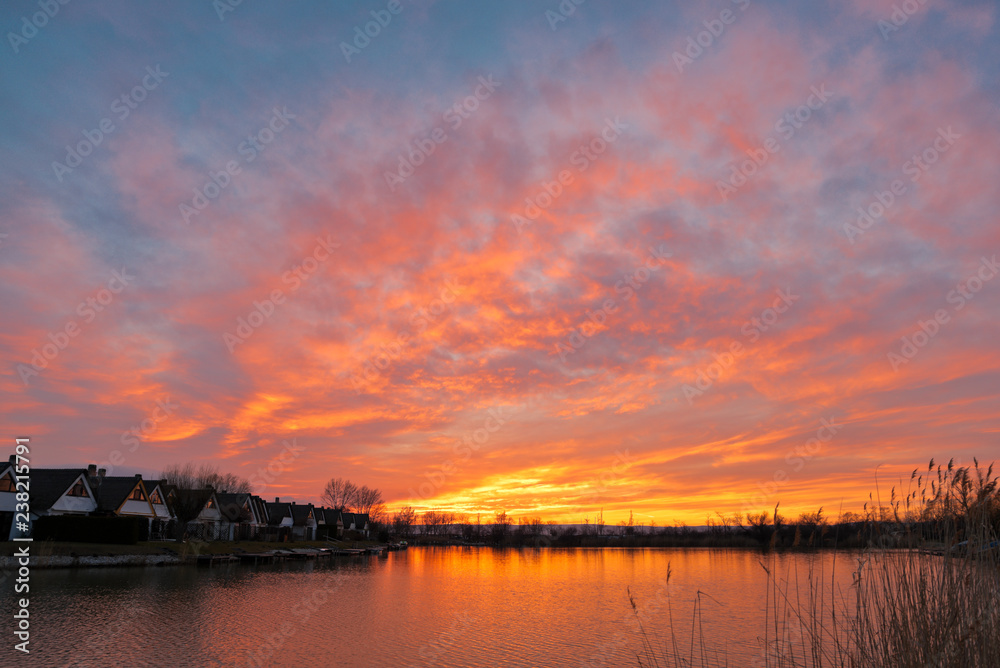Beautiful sunset at Lake Neusiedlersee in Austria with colorful orange and red illuminated cloudy sky. Picturesque holiday homes in terraced settlement. Reed grows on the shore.